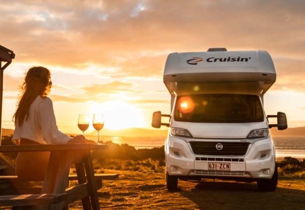 Eine Frau sitzt auf einer Bank neben ihrem Wohnmobil in Australien und schaut in den Sonnenuntergang. Auf dem Tisch neben ihr stehen zwei mit Rotwein gefüllte Weingläser./ Photo: © CRUISIN Motorhomes Eine Frau sitzt auf einer Bank neben ihrem Wohnmobil in Australien und schaut in den Sonnenuntergang. Auf dem Tisch neben ihr stehen zwei mit Rotwein gefüllte Weingläser.