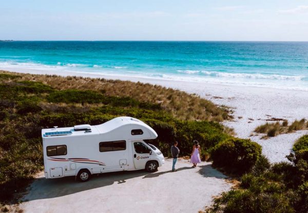 Ein Wohnmobil steht in Australien auf einem Stellplatz an der australischen Küste./ ©Photo: CRUISIN Motorhomes Ein Pärchen steht vor seinem Wohnmobil in Australien auf einem Stellplatz zwischen den Dünen an der australischen Küste. Hinter den Dünen sieht der Betrachter den Strand der ins türkisblaue übergeht.