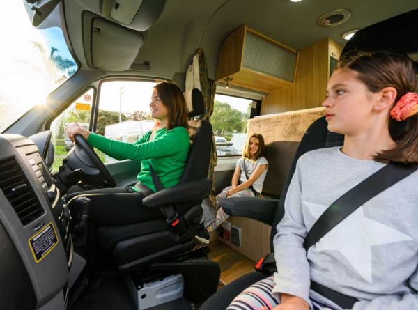 Eine Mutter am Steuer eines Campervans. Auf dem Beifahrersitz ein größeres Kind, auf dem hinteren Sitz ein jüngeres Mädchen.