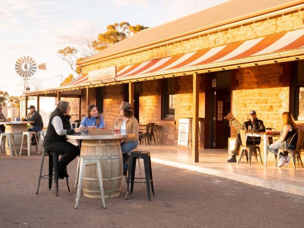 Prairie Hotel in den Flinders Ranges Südaustralien / ©Photo: Tourism Australia / South Australian Tourism Commission Menschen sitzen auf Barhockern an Stehtischen vor dem Prairie Hotel in den Flinders Ranges in Südaustralien