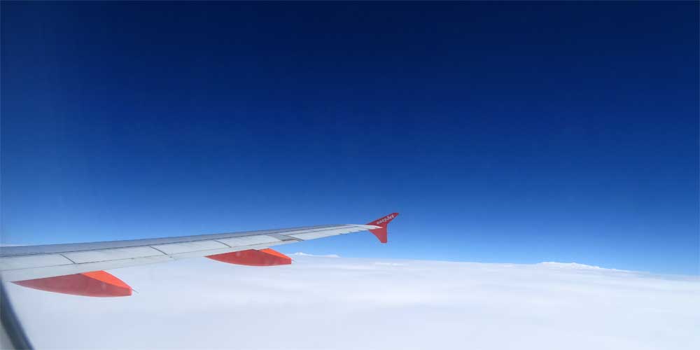 Aus einem Flugzeug sieht man den rechten Flügel genau an der Grenze zwischen einer weißen Wolkendecke und dem blauen Himmel