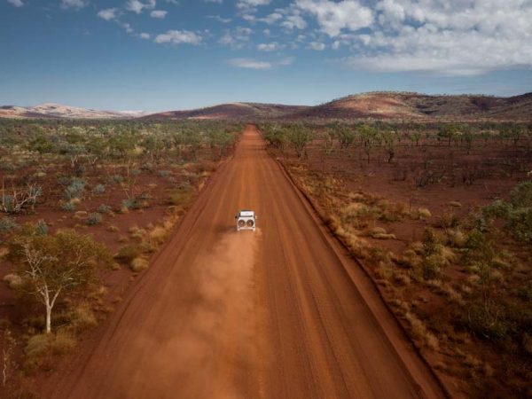 Ein Geländewagen auf einer Schotterstraße fährt auf den Horizont zu und zieht eine rote Staubwolke hinter sich her./ ©Photo: RedSands Camper Ein RedSands Geländewagen auf einer Schotterstraße aus der Sicht einer Drohne, fährt auf den Horizont zu und zieht eine rote Staubwolke hinter sich her.