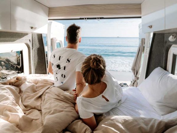 Zwei Menschen in Bett vor der geöffneten Doppeltür im Heck eines Campervans. Die Sonne scheint in das Fahrzeug/ Foto ©Cruisin Motorhomes Zwei Menschen in Bett vor der geöffneten Doppeltür im Heck eines Campervans. Die Sonne scheint in das Fahrzeug