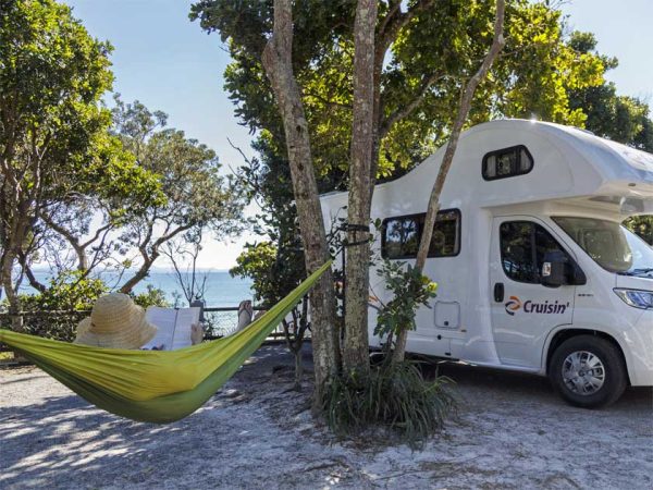 Eine Frau mit einem Strohhut liegt entspannt in einer Hängematte vor ihrem Camper und liest ein Buch/ ©Photo: Cruisin Motorhomes Eine Frau mit einem Strohhut liegt entspannt in einer Hängematte vor ihrem Camper und liest ein Buch